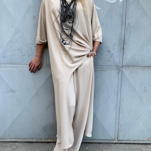 May include: A beige two-piece outfit consisting of a loose-fitting tunic with elbow-length sleeves and matching wide-leg trousers. The tunic features an asymmetrical hemline and is paired with a long, multi-layered black cord necklace.
