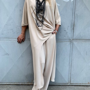 May include: A beige, flowing outfit consisting of a long tunic and wide-leg pants. A black, layered necklace adds contrast. The outfit has a relaxed fit and is made of a soft, draping fabric. The model is standing in front of a gray metal door.