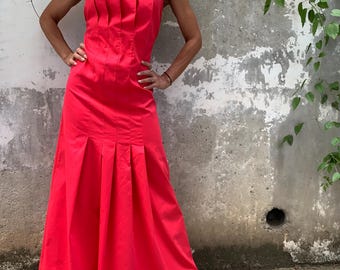 Puede incluir: Un vestido rojo vibrante con un corpiño plisado y una falda fluida. El vestido presenta un escote cuadrado y tirantes negros. La modelo lleva sandalias de plataforma negras. El fondo es una pared gris texturizada con follaje verde.
