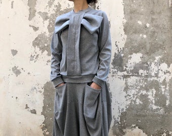 Könnte beinhalten: Ein graues Sweatshirt mit einer großen Schleife am Ausschnitt und einem passenden grauen Rock. Der Rock hat ein gerafftes Design und große Taschen an den Seiten. Das Model trägt weiße Sneaker mit silbernen Akzenten.