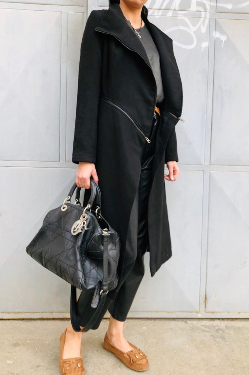 May include: A black long coat with a zipper detail and a black leather handbag. The person is wearing brown suede loafers.