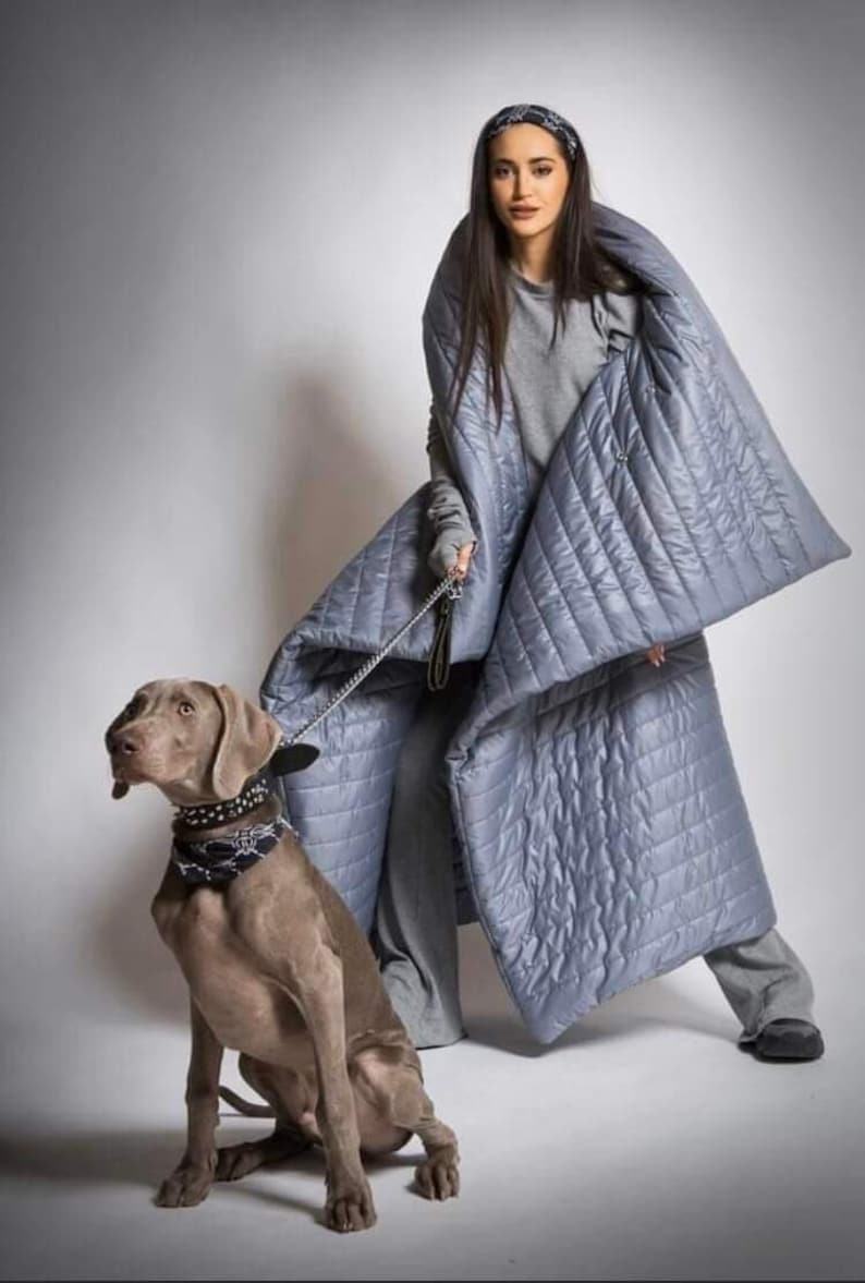 May include: A person wearing a quilted, light blue wrap and matching pants, holding a leash attached to a Weimaraner dog. The dog wears a black collar with white accents. The wrap has a diamond pattern.