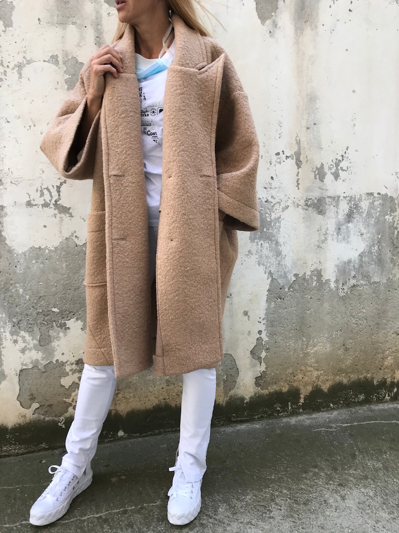 Może przedstawiać: Długi, brązowy płaszcz wełniany z szerokim kołnierzem i dużymi kieszeniami. Płaszcz jest rozpięty, a rękawy podwinięte. Osoba nosząca płaszcz stoi przed szarą ścianą.