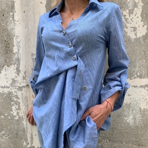 May include: A light blue button-down shirt with a unique asymmetrical design. The shirt features long sleeves that can be rolled up, a collared neckline, and a wrap-style front secured with buttons. The fabric has a textured appearance.