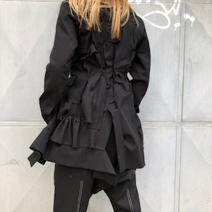 Puede incluir: Una chaqueta negra con un dobladillo con volantes y un diseño único en la espalda. La chaqueta está hecha de una tela suave y texturizada.