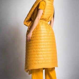 May include: A mustard yellow quilted ensemble consisting of a vest, long skirt, and wide-leg pants. The outfit is worn over a yellow t-shirt. The model is facing away from the camera, with dark hair.