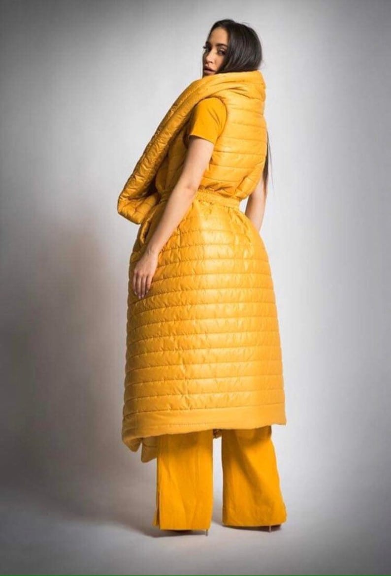 May include: A mustard yellow quilted ensemble consisting of a vest, long skirt, and wide-leg pants. The outfit is worn over a yellow t-shirt. The model is facing away from the camera, with dark hair.