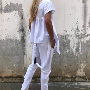 Puede incluir: Un pantal&oacute;n de traje de lino blanco con una parte superior holgada y pantalones de pierna ancha. La parte superior tiene un dobladillo con volantes y los pantalones tienen un detalle de correa negra.