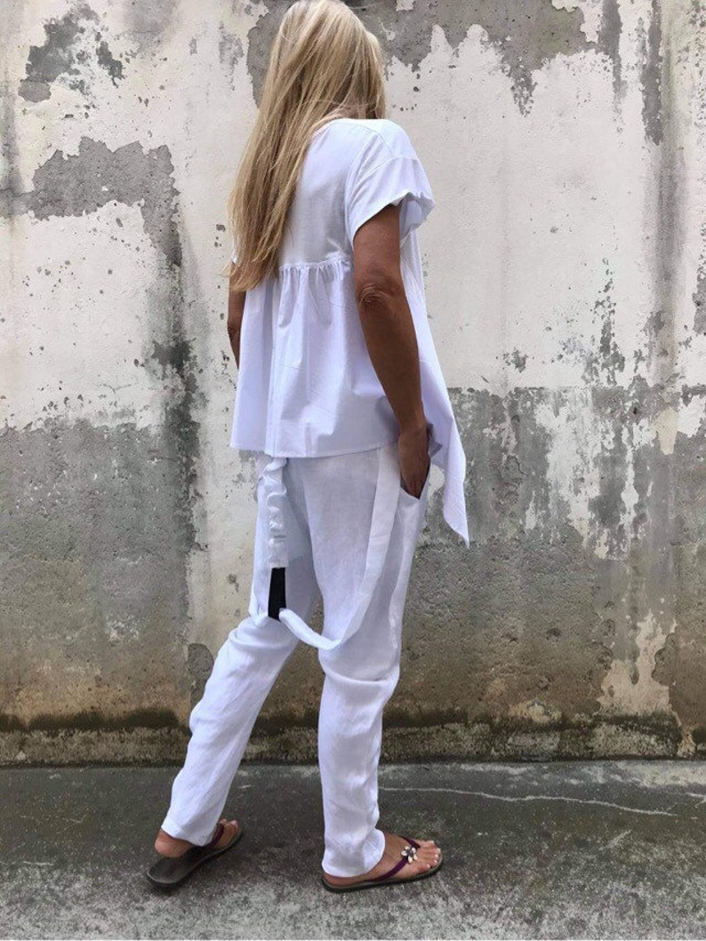 Puede incluir: Un pantal&oacute;n de traje de lino blanco con una parte superior holgada y pantalones de pierna ancha. La parte superior tiene un dobladillo con volantes y los pantalones tienen un detalle de correa negra.