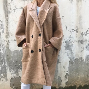 Może przedstawiać: Długi, dwurzędowy płaszcz w kolorze camel z dużymi kieszeniami. Płaszcz wykonany jest z miękkiej, puszystej tkaniny.