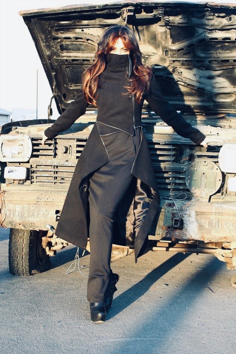 May include: A woman wearing a black long-sleeved coat with a high collar and a long, flowing skirt. The coat has a zipper closure and a belt. The woman is standing in front of a vintage car with the hood open.