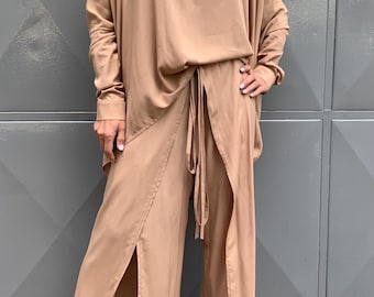 May include: A two-piece outfit in a light brown color. The top has a collared neckline and long sleeves. The pants have a wide leg and a slit on each side.