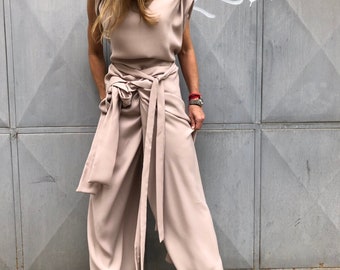 May include: A woman wearing a light brown jumpsuit with a wide leg and a sash tied at the waist. The jumpsuit has a one-shoulder neckline and short sleeves. She is wearing nude heels.