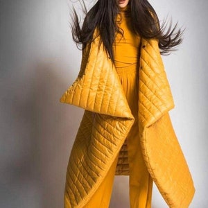 May include: A woman wearing a mustard yellow quilted coat and matching pants. The coat has a unique design with large, angular sleeves. The outfit is paired with a mustard yellow turtleneck. The background is a solid white.