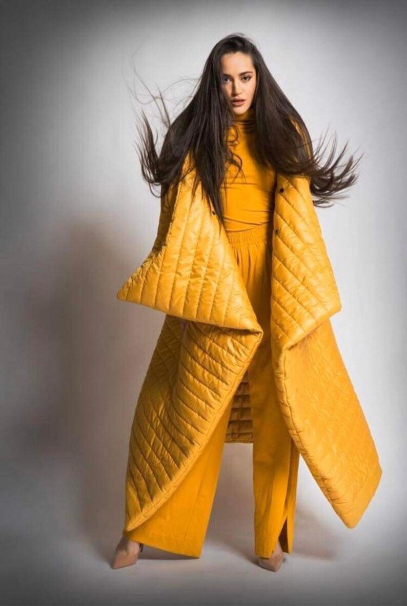 May include: A woman wearing a mustard yellow quilted coat and matching pants. The coat has a unique design with large, angular sleeves. The outfit is paired with a mustard yellow turtleneck. The background is a solid white.