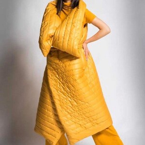 May include: A mustard yellow quilted coat and matching pants. The coat is long and wraps around the body, with a high collar. The pants are wide-legged. The outfit is worn with a yellow short-sleeved shirt and nude heels.