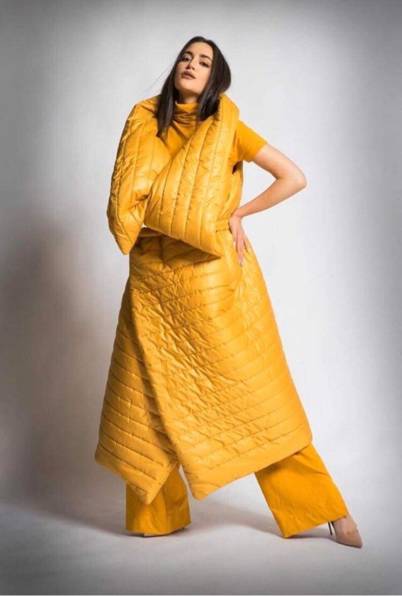 May include: A mustard yellow quilted coat and matching pants. The coat is long and wraps around the body, with a high collar. The pants are wide-legged. The outfit is worn with a yellow short-sleeved shirt and nude heels.