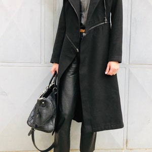 May include: A black long coat with a zipper closure and a long, flowing silhouette. The coat has a collar and is worn over a black top and black pants. The person is wearing brown suede moccasins.