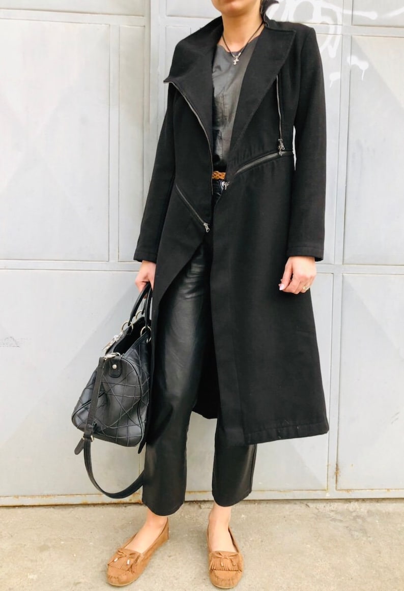 May include: A black long coat with a zipper closure and a long, flowing silhouette. The coat has a collar and is worn over a black top and black pants. The person is wearing brown suede moccasins.