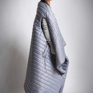 May include: A light gray, quilted, oversized blanket with a diagonal stitch pattern. The blanket has arm openings and a hood. The person is wearing a gray long-sleeved shirt. The blanket is designed to be worn.