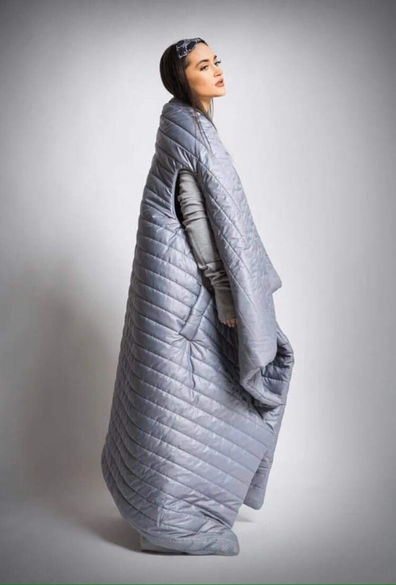 May include: A light gray, quilted, oversized blanket with a diagonal stitch pattern. The blanket has arm openings and a hood. The person is wearing a gray long-sleeved shirt. The blanket is designed to be worn.