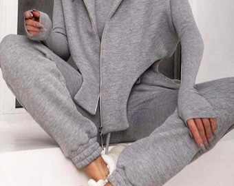 Könnte beinhalten: Ein grauer zweiteiliger Trainingsanzug mit einer Reißverschlussjacke mit übertriebenen Schultern und dem Wort "Momenter" auf der Vorderseite. Das Model trägt weiße Turnschuhe.