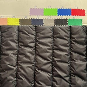 May include: Close-up of a black quilted fabric with vertical stitching. Above the fabric are color swatches in various shades, including pink, yellow, blue, green, and purple. The fabric has a shiny, smooth texture.
