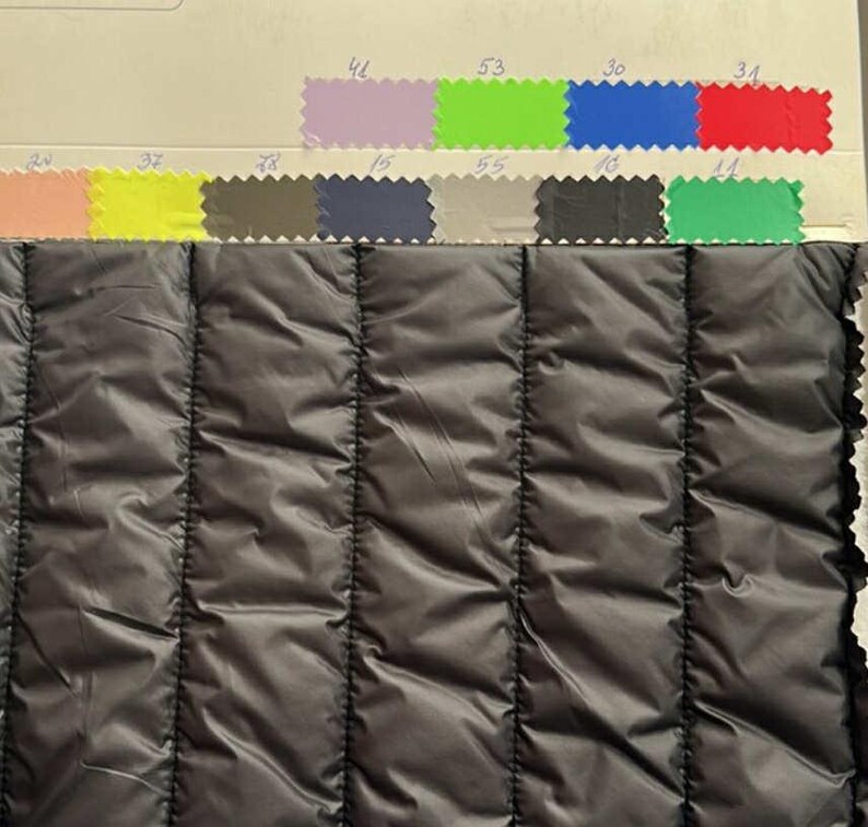 May include: Close-up of a black quilted fabric with vertical stitching. Above the fabric are color swatches in various shades, including pink, yellow, blue, green, and purple. The fabric has a shiny, smooth texture.