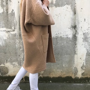 Może przedstawiać: Długi, brązowy, puszysty płaszcz z dużą kieszenią po lewej stronie. Płaszcz jest noszony na białych spodniach i białych butach sportowych.