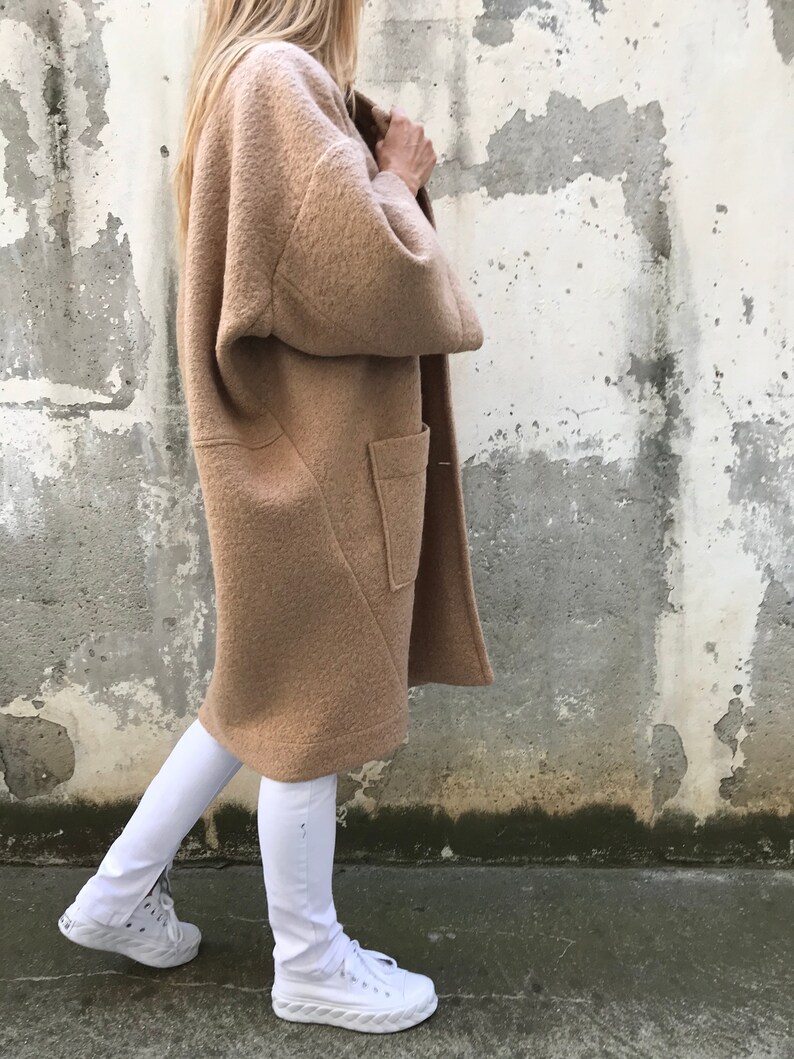 Może przedstawiać: Długi, brązowy, puszysty płaszcz z dużą kieszenią po lewej stronie. Płaszcz jest noszony na białych spodniach i białych butach sportowych.