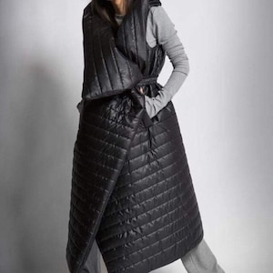 May include: A black quilted wrap coat with a hood and belt. The coat is long, reaching the ankles, and has a diagonal wrap design. The model is wearing a gray long-sleeved shirt and gray pants.