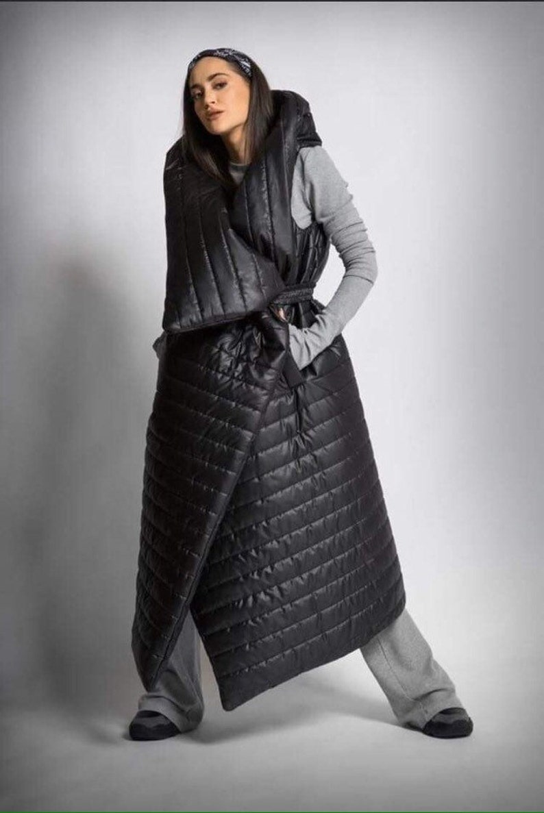 May include: A black quilted wrap coat with a hood and belt. The coat is long, reaching the ankles, and has a diagonal wrap design. The model is wearing a gray long-sleeved shirt and gray pants.