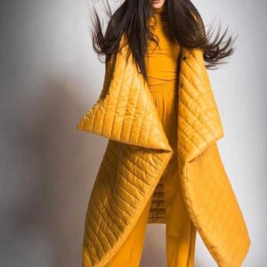 May include: A woman wearing a mustard yellow quilted coat and matching pants. The coat has large, angular sleeves and a diamond pattern. The outfit is a monochromatic ensemble, creating a bold fashion statement.