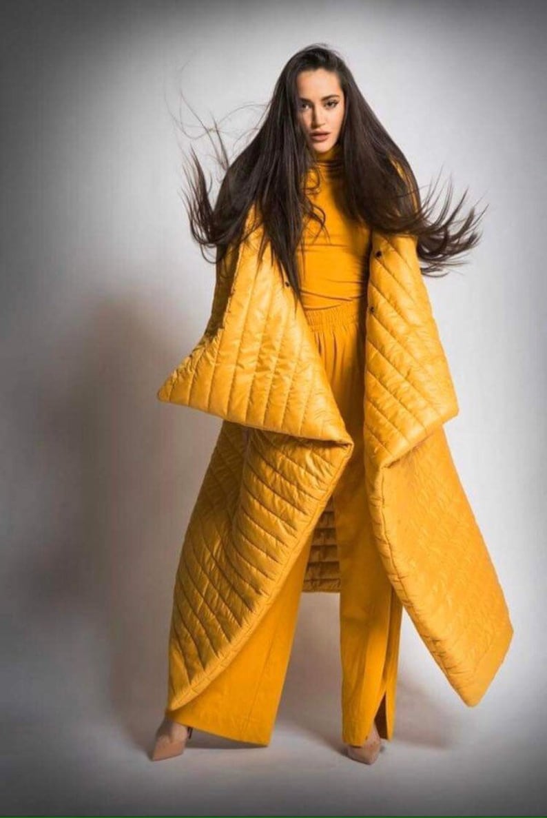 May include: A woman wearing a mustard yellow quilted coat and matching pants. The coat has large, angular sleeves and a diamond pattern. The outfit is a monochromatic ensemble, creating a bold fashion statement.