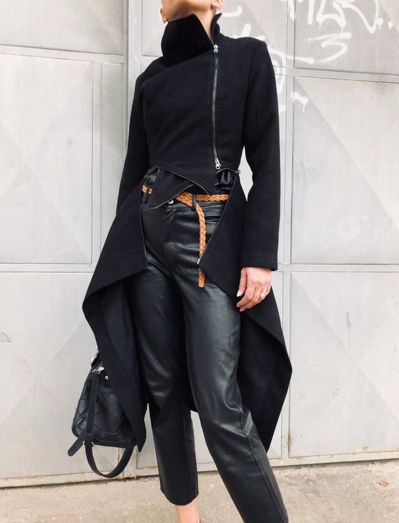 May include: A black asymmetrical jacket with a high collar and a zipper closure. The jacket has a long, flowing back panel and a shorter front panel. The jacket is worn with a brown leather belt and black leather pants.