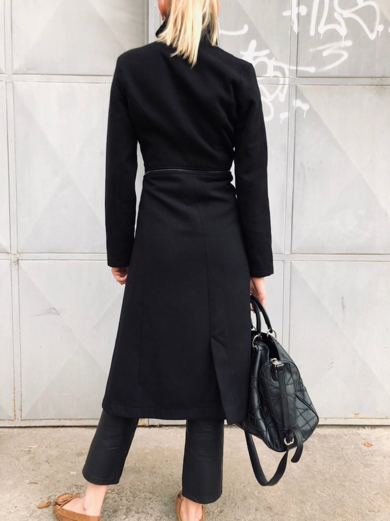 May include: A long black coat with a high collar and a belt. The coat is made of a thick fabric and has a simple design. The model is wearing black pants and brown shoes.