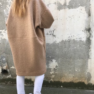 Może przedstawiać: Długi, brązowy, oversize'owy płaszcz wełniany o luźnym kroju. Płaszcz ma prosty d&oacute;ł i dwie duże kieszenie po bokach. Modelka ma na sobie białe spodnie i białe buty sportowe.