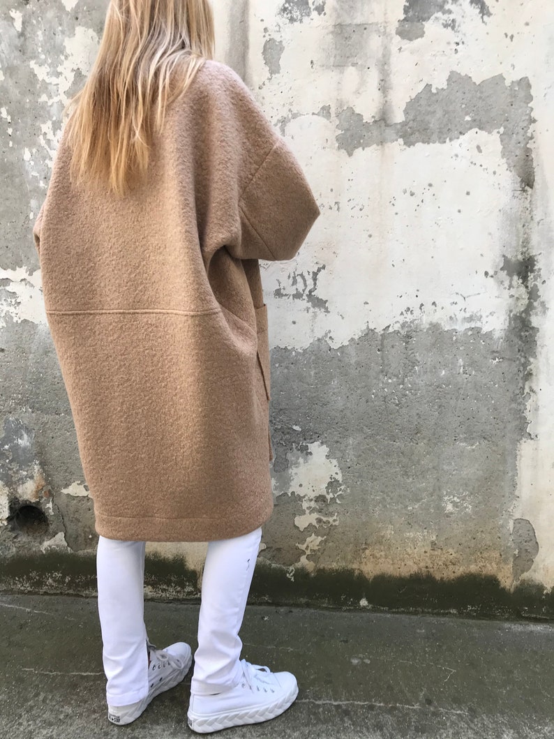 Może przedstawiać: Długi, brązowy, oversize'owy płaszcz wełniany o luźnym kroju. Płaszcz ma prosty d&oacute;ł i dwie duże kieszenie po bokach. Modelka ma na sobie białe spodnie i białe buty sportowe.