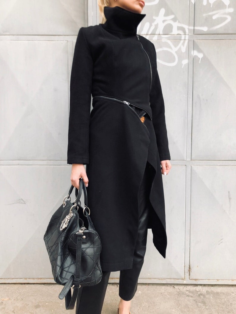 May include: A black long-sleeved coat with a high collar and a zipper closure. The coat has a diagonal zipper detail and a belt. The person is wearing black pants and carrying a black leather handbag.