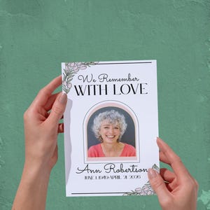 Può includere: Un biglietto commemorativo tenuto in mano, con la foto di una donna sorridente all'interno di una cornice ad arco. Il biglietto recita "We Remember WITH LOVE" e "Ann Robertson" con date di nascita e di morte. Accenti floreali adornano i bordi.