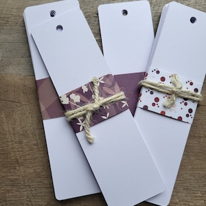 Puede incluir: Tres pilas de tarjetas de marcadores blancas con agujeros perforados en la parte superior. Las pilas est&aacute;n atadas con hilo y decoradas con papel estampado. El papel de la pila de la izquierda tiene un fondo morado con dise&ntilde;os florales blancos. El papel de la pila de la derecha tiene un fondo blanco con lunares rojos y marrones.
