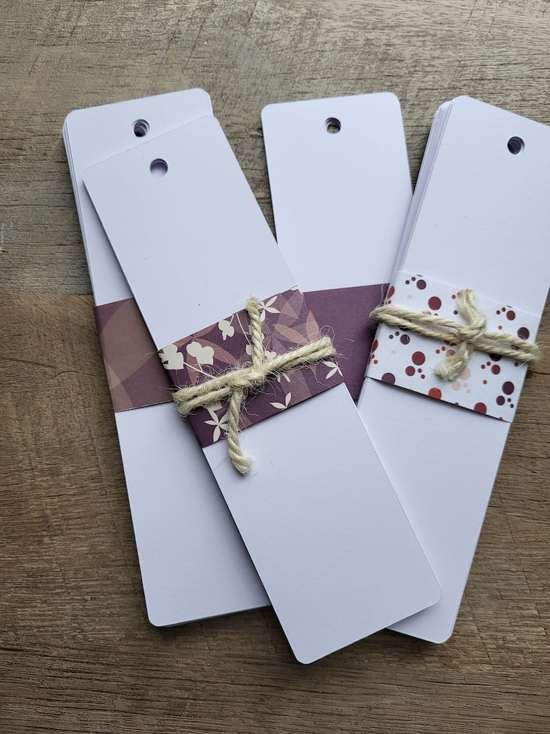 Puede incluir: Tres pilas de tarjetas de marcadores blancas con agujeros perforados en la parte superior. Las pilas est&aacute;n atadas con hilo y decoradas con papel estampado. El papel de la pila de la izquierda tiene un fondo morado con dise&ntilde;os florales blancos. El papel de la pila de la derecha tiene un fondo blanco con lunares rojos y marrones.