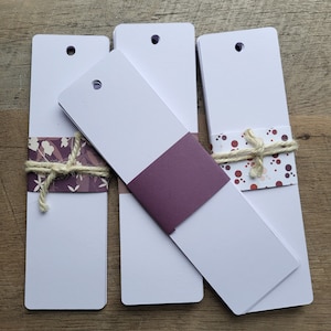 Puede incluir: Tres pilas de marcadores de libros blancos en blanco con un agujero perforado en la parte superior. El marcador superior de cada pila tiene un dise&ntilde;o de papel decorativo. Un marcador tiene un dise&ntilde;o floral morado y blanco, otro tiene un color morado s&oacute;lido y el &uacute;ltimo tiene un dise&ntilde;o de lunares rosa y blanco.