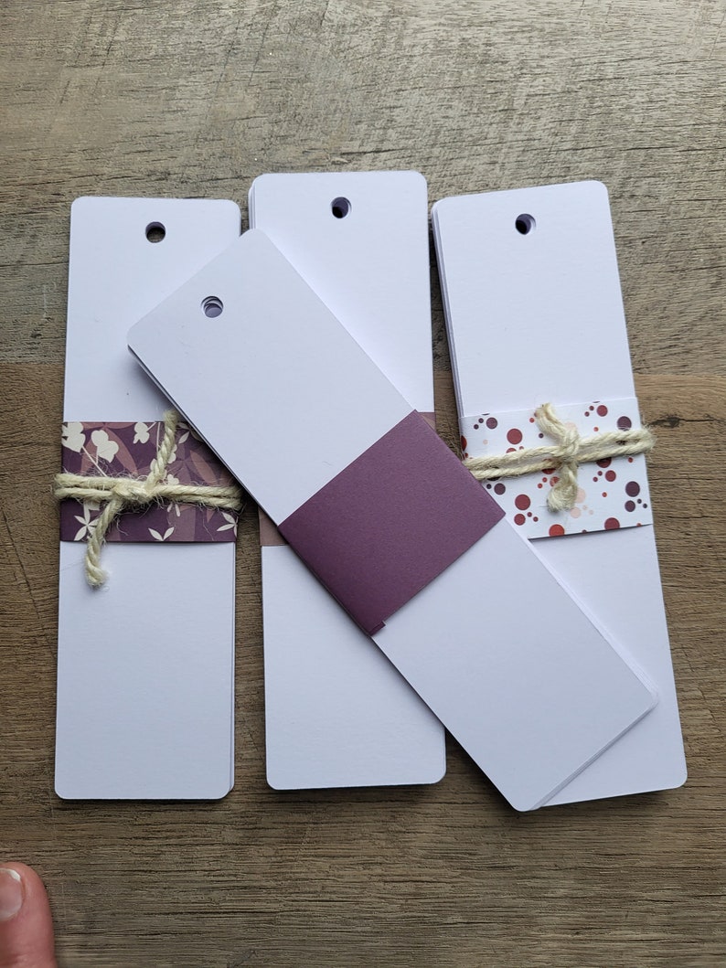 Puede incluir: Tres pilas de marcadores de libros blancos en blanco con un agujero perforado en la parte superior. El marcador superior de cada pila tiene un dise&ntilde;o de papel decorativo. Un marcador tiene un dise&ntilde;o floral morado y blanco, otro tiene un color morado s&oacute;lido y el &uacute;ltimo tiene un dise&ntilde;o de lunares rosa y blanco.