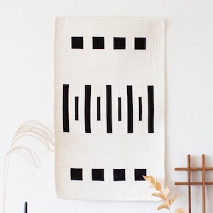 Op de afbeelding: Een rechthoekig wandkleed van gebroken wit textiel met een geometrisch patroon van zwarte vierkanten en verticale balken. Het item hangt boven een houten dressoir met messing handgrepen, naast een houten rasterframe en decoratie.