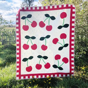 Puede incluir: Una colcha rectangular con fondo color crema y un patrón repetitivo de cerezas rojas, tallos verdes y hojas de color verde oscuro. La colcha está enmarcada por un borde a cuadros rojos y blancos con un remate exterior rojo.