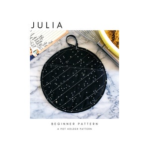 May include: A black and white pot holder with a star pattern. The pot holder has a pocket on the side and a loop for hanging. The text "BEGINNER PATTERN" and "A POT HOLDER PATTERN" is printed below the pot holder.