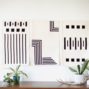 May include: Three black and white geometric wall hangings with abstract designs. The hangings are on a white wall above a wooden table with plants and antlers.