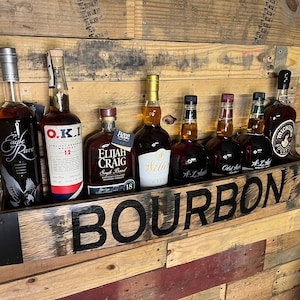 Pu&ograve; includere: Un ripiano in legno per bottiglie di bourbon con la scritta "BOURBON" in lettere nere. Il ripiano &egrave; pieno di varie bottiglie di bourbon, tra cui Eagle Rare, O.K.I., Elijah Craig, Weller, W.L. Weller, Old Forester e Michter's.