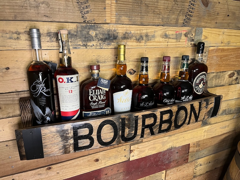 Pu&ograve; includere: Un ripiano in legno per bottiglie di bourbon con la scritta "BOURBON" in lettere nere. Il ripiano &egrave; pieno di varie bottiglie di bourbon, tra cui Eagle Rare, O.K.I., Elijah Craig, Weller, W.L. Weller, Old Forester e Michter's.